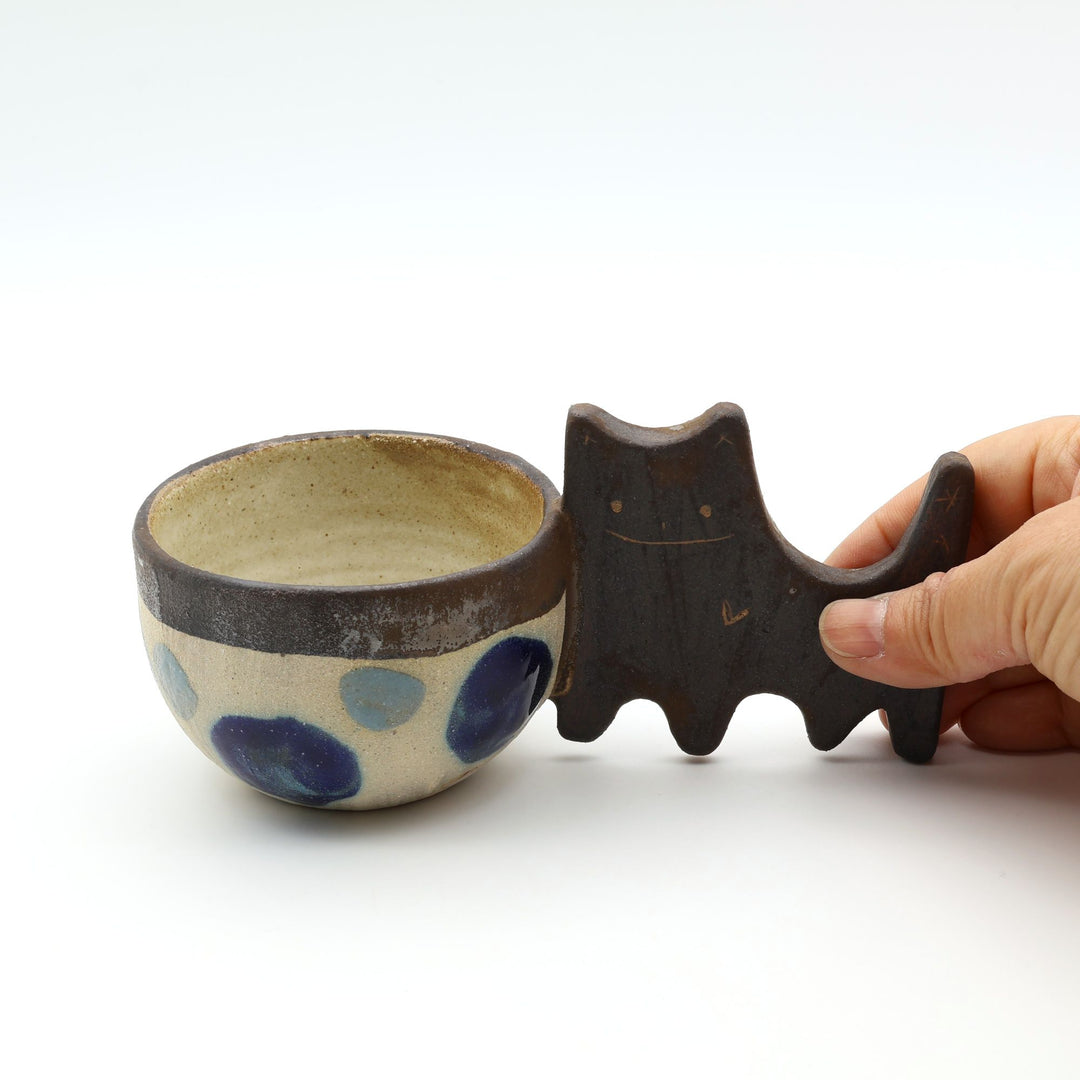 沖縄のやちむん「土の種」によるネコをモチーフにした四つ足カップ。ドット柄のカップに、ネコの体と足を模したユニークな持ち手が組み合わさった、遊び心あふれる陶器のマグカップ。- canosaオンラインショップ