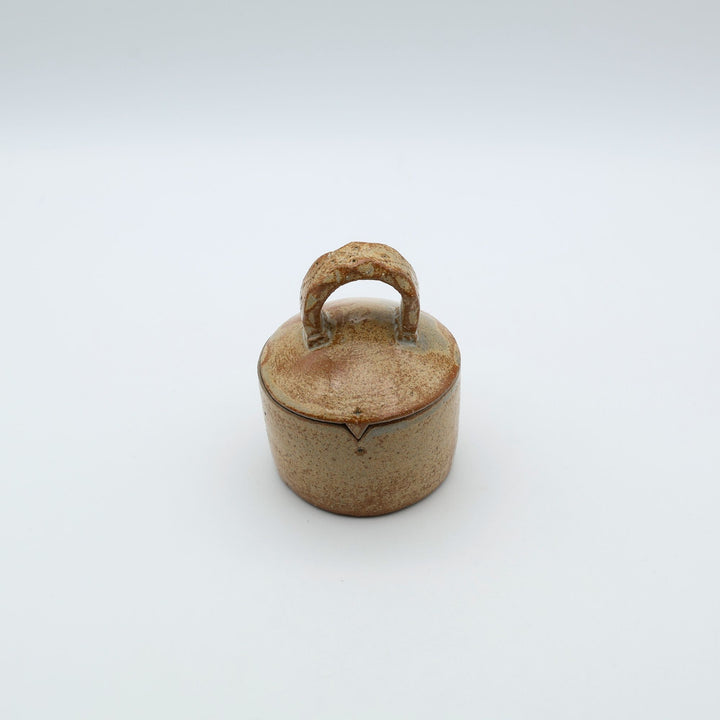 沖縄のやちむん工房「土の種」による素朴な土の質感の陶器の蓋物（キャニスター）。大きな持ち手と蓋のズレを防ぐ突起が特徴的な、調味料やアクセサリー入れに最適な手仕事のうつわ。