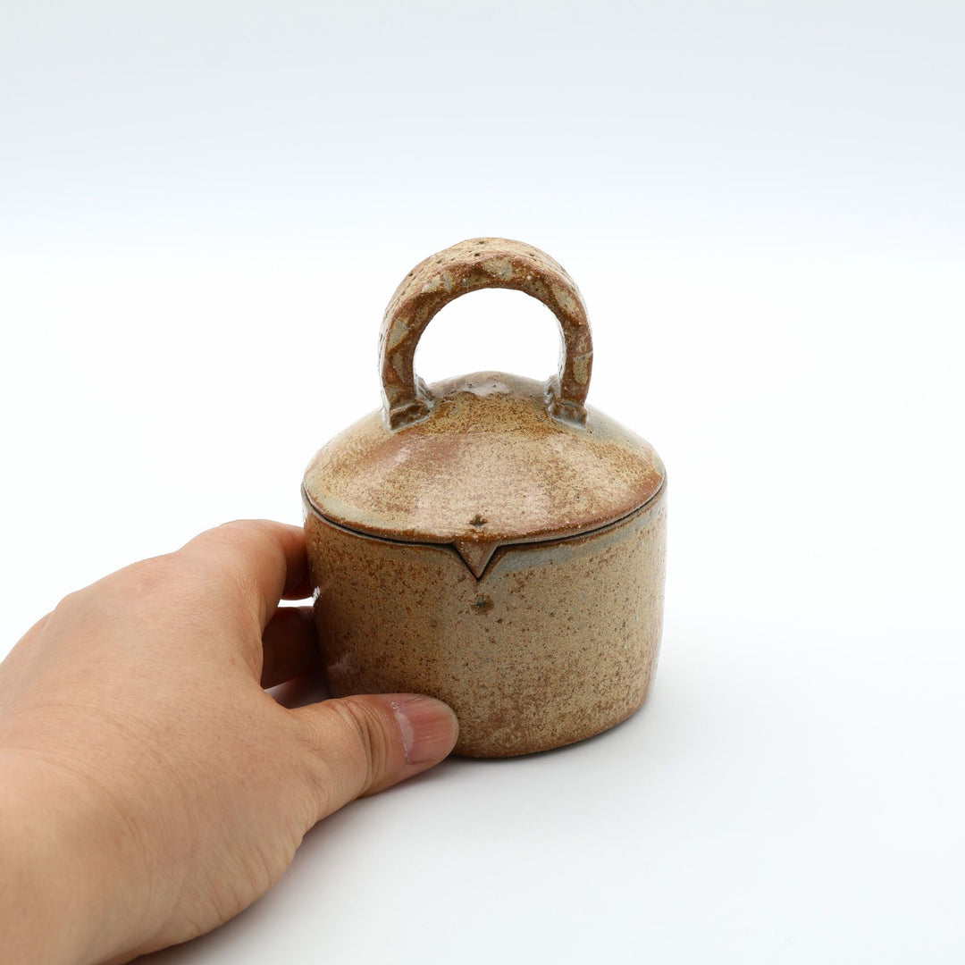 沖縄のやちむん工房「土の種」による素朴な土の質感の陶器の蓋物（キャニスター）。大きな持ち手と蓋のズレを防ぐ突起が特徴的な、調味料やアクセサリー入れに最適な手仕事のうつわ。