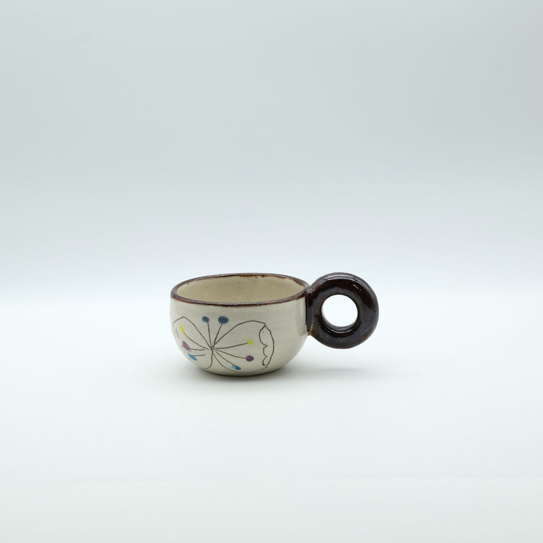 うるま市のやちむん工房「土の種」作、外側にお花と蝶々（ちょうちょ）が描かれた粉引きマグカップ。丸い茶色のリング型持ち手が特徴。/ Handcrafted Kohiki ceramic mug with whimsical flower and butterfly illustrations and a distinct circular handle by Tsuchi no Tane, Okinawa.