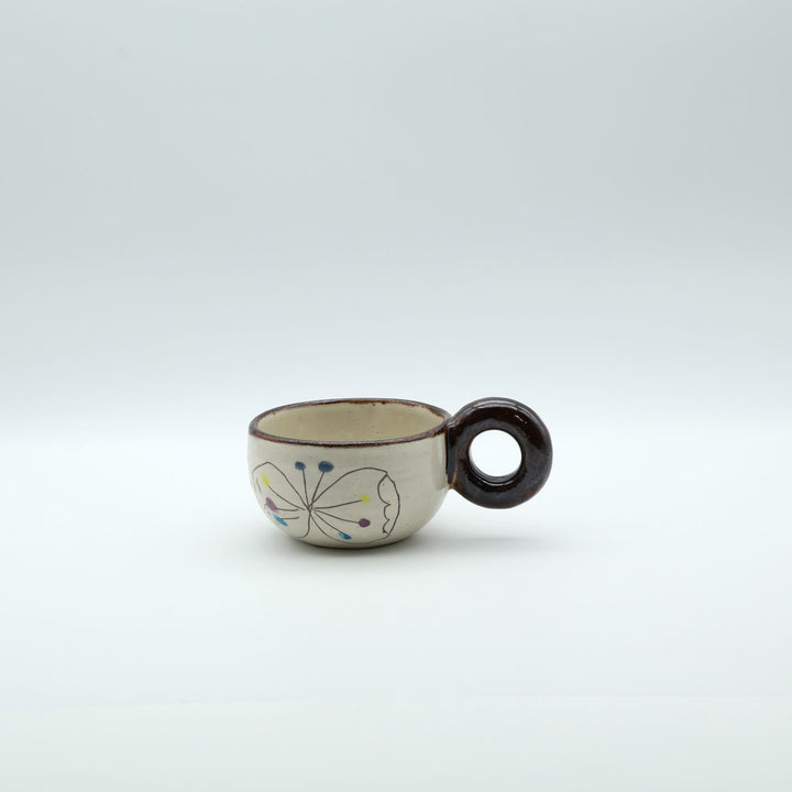 うるま市のやちむん工房「土の種」作、外側にお花と蝶々（ちょうちょ）が描かれた粉引きマグカップ。丸い茶色のリング型持ち手が特徴。/ Handcrafted Kohiki ceramic mug with whimsical flower and butterfly illustrations and a distinct circular handle by Tsuchi no Tane, Okinawa.