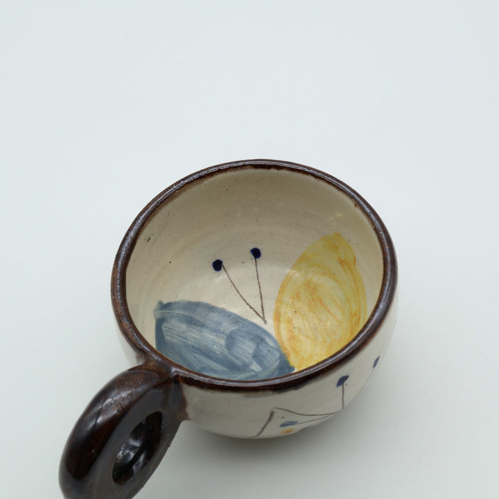 沖縄の陶器「土の種」の粉引マグカップ。器の内側に大胆に描かれた大きな青と黄色の蝶々の絵付けと、しっかり握れる丸い持ち手。/ Inside view of an Okinawan pottery mug by Tsuchi no Tane, featuring a large, beautiful blue and yellow butterfly motif painted on the interior.