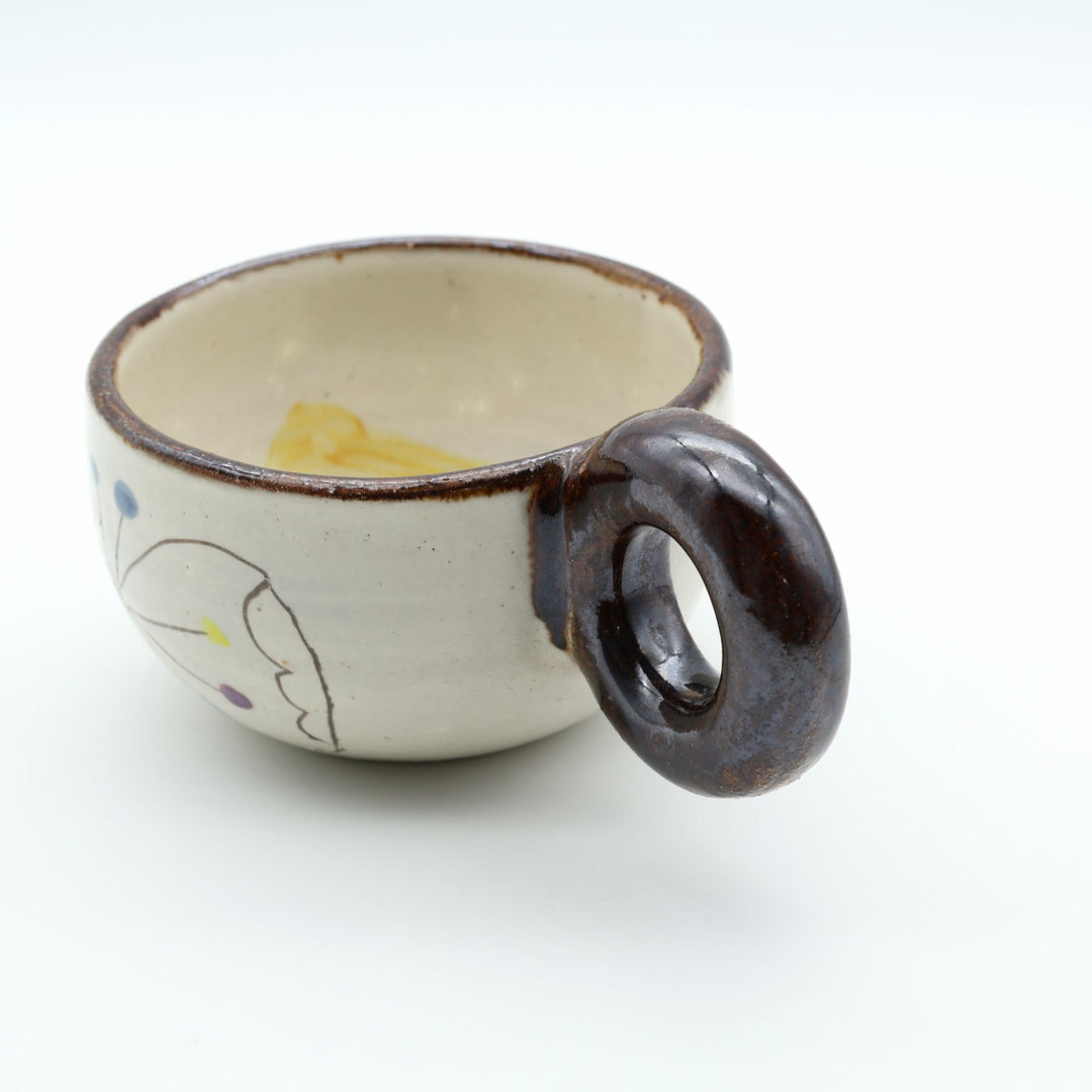 うるま市のやちむん工房「土の種」作、外側にお花と蝶々（ちょうちょ）が描かれた粉引きマグカップ。丸い茶色のリング型持ち手が特徴。/ Handcrafted Kohiki ceramic mug with whimsical flower and butterfly illustrations and a distinct circular handle by Tsuchi no Tane, Okinawa.