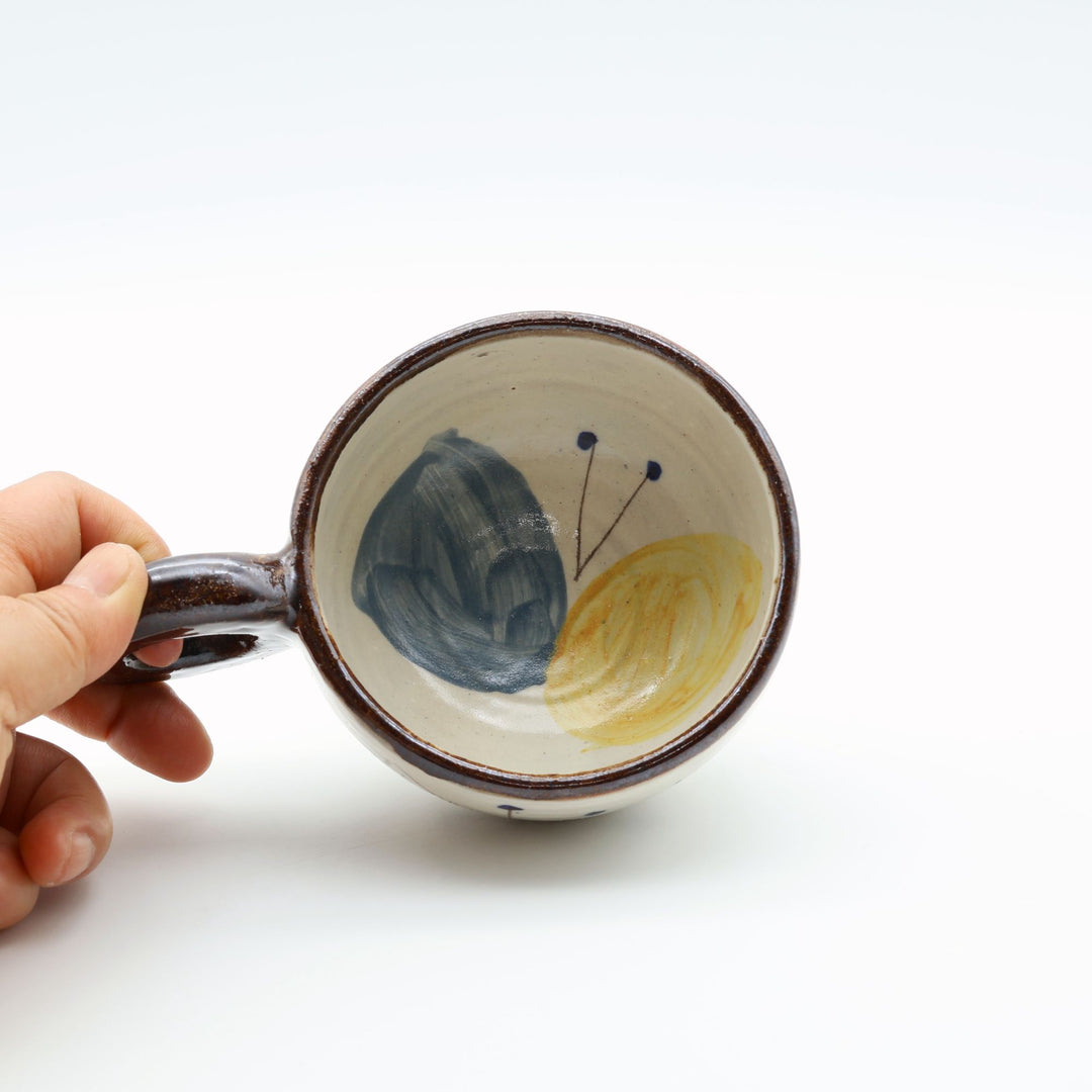 沖縄の陶器「土の種」の粉引マグカップ。器の内側に大胆に描かれた大きな青と黄色の蝶々の絵付けと、しっかり握れる丸い持ち手。/ Inside view of an Okinawan pottery mug by Tsuchi no Tane, featuring a large, beautiful blue and yellow butterfly motif painted on the interior.