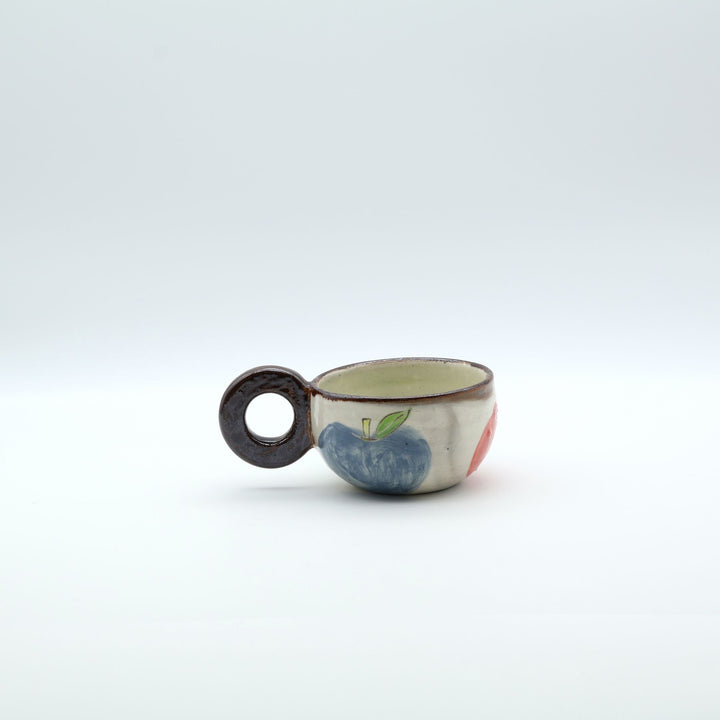 沖縄の陶器「土の種」のリンゴ柄粉引マグカップ。外側に並ぶ3色のリンゴと、内側にワンポイントで描かれた青リンゴ。/ Artisan Okinawan pottery mug by Tsuchi no Tane, showing the colorful apple motifs on the outside and a single green apple painted inside.