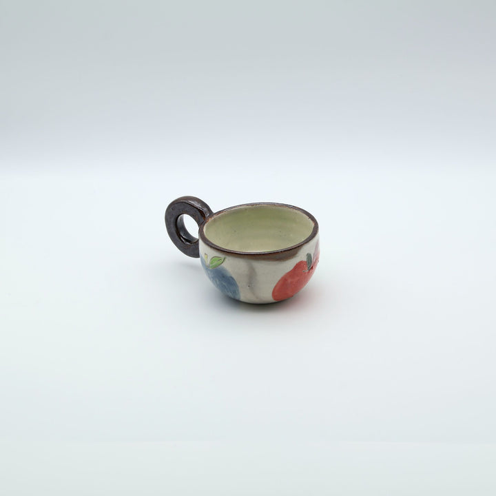 沖縄の陶器「土の種」のリンゴ柄粉引マグカップ。外側に並ぶ3色のリンゴと、内側にワンポイントで描かれた青リンゴ。/ Artisan Okinawan pottery mug by Tsuchi no Tane, showing the colorful apple motifs on the outside and a single green apple painted inside.