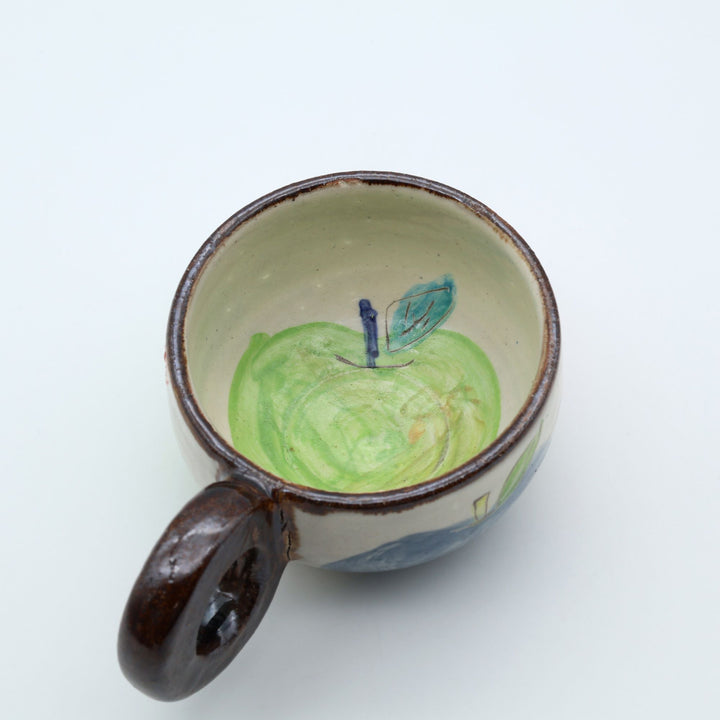 沖縄の陶器「土の種」のリンゴ柄粉引マグカップ。外側に並ぶ3色のリンゴと、内側にワンポイントで描かれた青リンゴ。/ Artisan Okinawan pottery mug by Tsuchi no Tane, showing the colorful apple motifs on the outside and a single green apple painted inside.