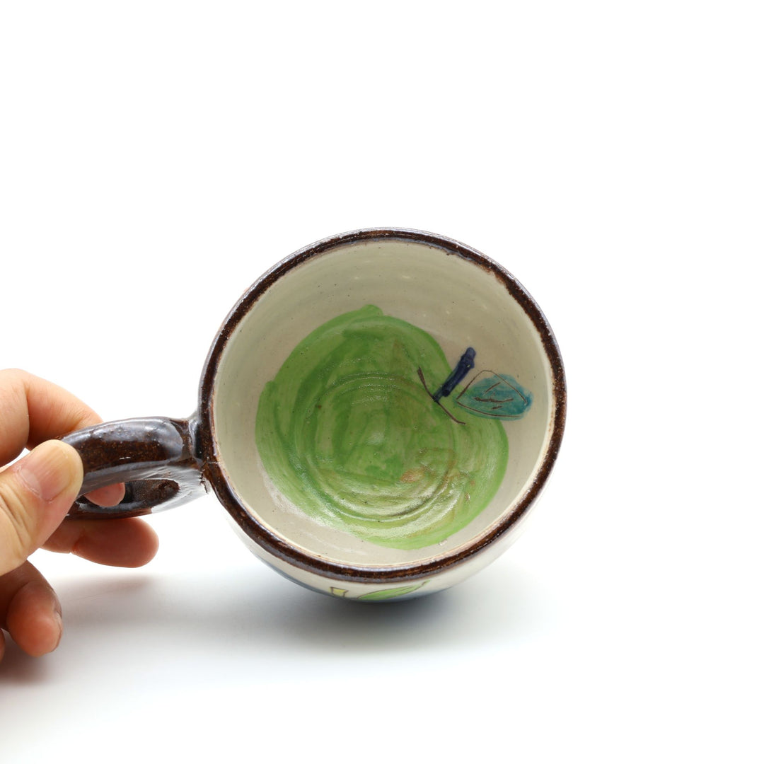 沖縄の陶器「土の種」のリンゴ柄粉引マグカップ。外側に並ぶ3色のリンゴと、内側にワンポイントで描かれた青リンゴ。/ Artisan Okinawan pottery mug by Tsuchi no Tane, showing the colorful apple motifs on the outside and a single green apple painted inside.