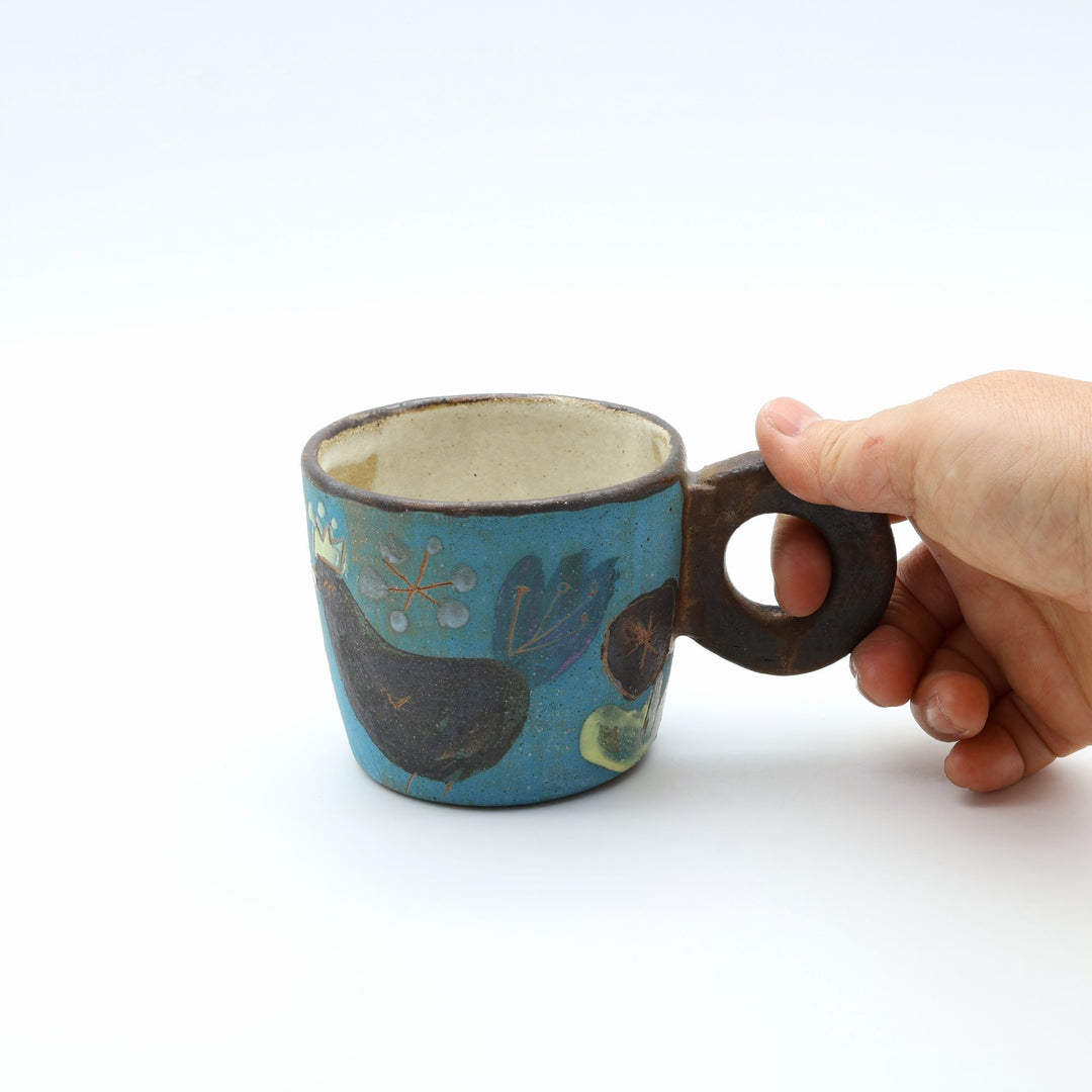 沖縄のやちむん工房「土の種」、鳥と花が描かれた青い1点ものマグカップ。ユニークな丸いリング型の持ち手を手が握っている様子。/ Handcrafted one-of-a-kind Okinawan Yachumun ceramic mug by Tsuchi no Tane, featuring a unique round ring handle and bird motif.