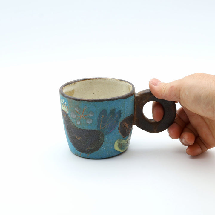 沖縄のやちむん工房「土の種」、鳥と花が描かれた青い1点ものマグカップ。ユニークな丸いリング型の持ち手を手が握っている様子。/ Handcrafted one-of-a-kind Okinawan Yachumun ceramic mug by Tsuchi no Tane, featuring a unique round ring handle and bird motif.