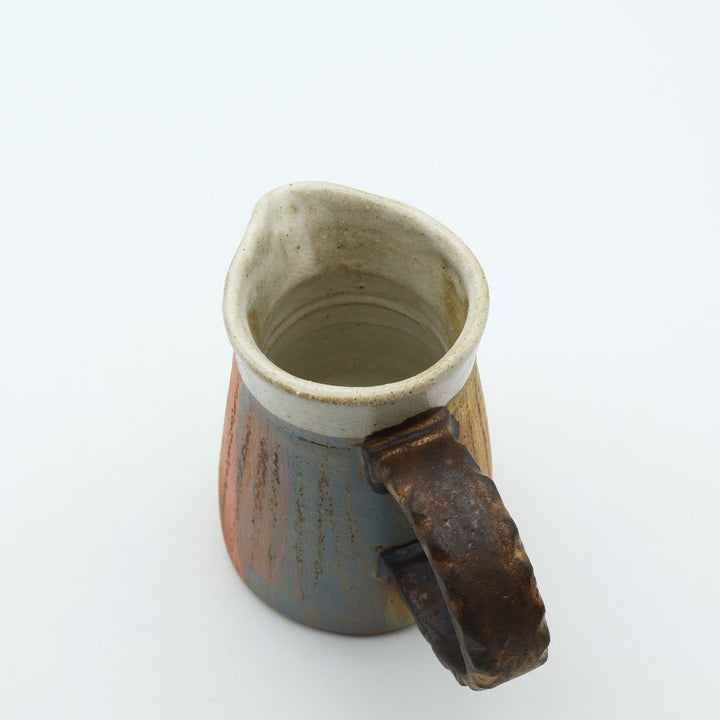 うるま市のやちむん工房「土の種」作、白化粧と虹色のグラデーションが美しい陶器のピッチャー。手彫りのギザギザとした彫理が施された持ち手。/ Close-up of a handcrafted Okinawan Yachumun ceramic pitcher by Tsuchi no Tane, featuring a hand-carved textured handle and intricate glaze work.