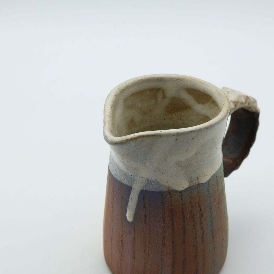 うるま市のやちむん工房「土の種」作、白化粧と緑・茶のグラデーションが美しい陶器のピッチャー。手彫りのギザギザとした彫理（しのぎ）が施された持ち手。/ Close-up of a handcrafted Okinawan Yachumun ceramic pitcher by Tsuchi no Tane, featuring a hand-carved textured handle and intricate glaze work.