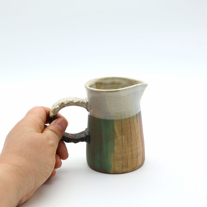 うるま市のやちむん工房「土の種」作、白化粧と緑・茶のグラデーションが美しい陶器のピッチャー。手彫りのギザギザとした彫理（しのぎ）が施された持ち手。/ Close-up of a handcrafted Okinawan Yachumun ceramic pitcher by Tsuchi no Tane, featuring a hand-carved textured handle and intricate glaze work.