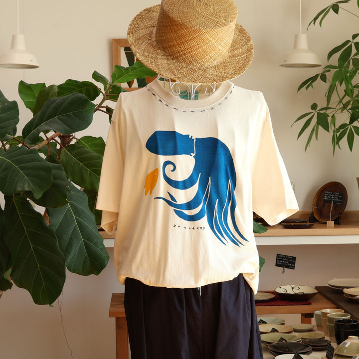 Doucatty（ドゥカティ）の手捺染Tシャツ「たここんにちは」。優しく温かみのある生成り地に、鮮やかなブルーで足をしなやかにくねらせる大きなタコがフロント中央に描かれ、足元には小さな黄色い魚があしらわれている。沖縄製。