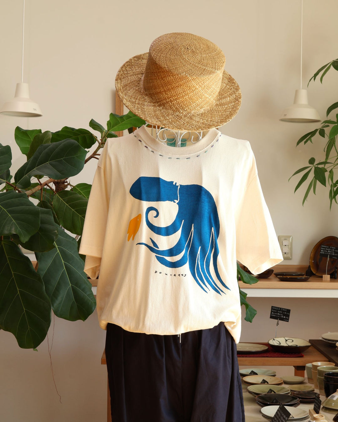 Doucatty（ドゥカティ）の手捺染Tシャツ「たここんにちは」。優しく温かみのある生成り地に、鮮やかなブルーで足をしなやかにくねらせる大きなタコがフロント中央に描かれ、足元には小さな黄色い魚があしらわれている。首元には青いステッチが。沖縄製。