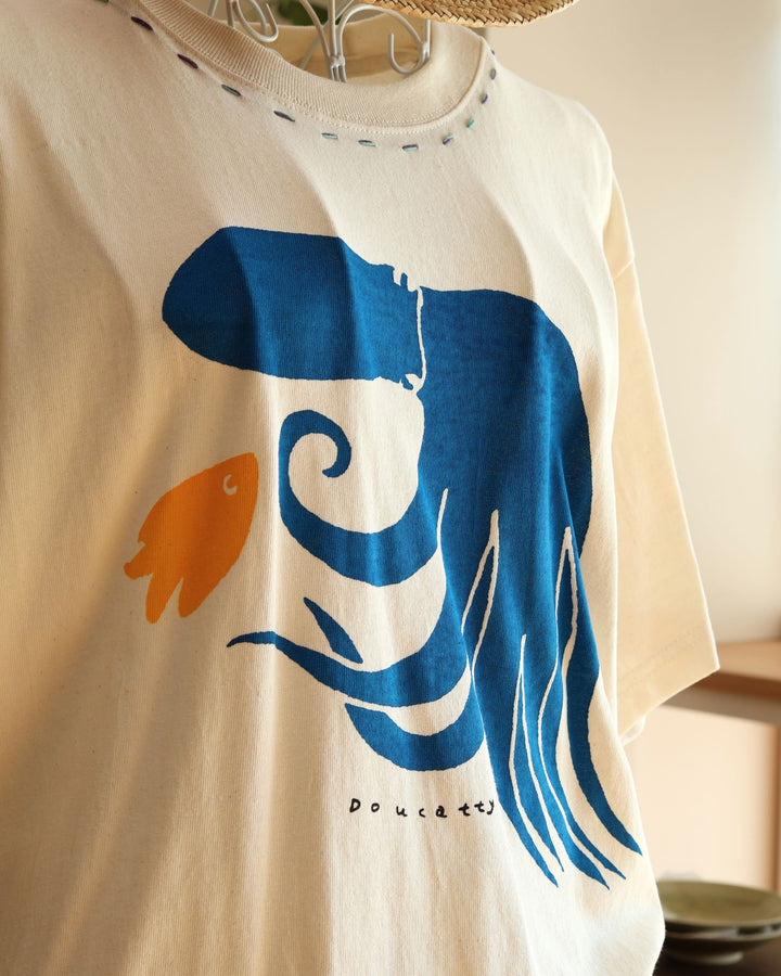 Doucatty（ドゥカティ）の手捺染Tシャツ「たここんにちは」。優しく温かみのある生成り地に、鮮やかなブルーで足をしなやかにくねらせる大きなタコがフロント中央に描かれ、足元には小さな黄色い魚があしらわれている。沖縄製。