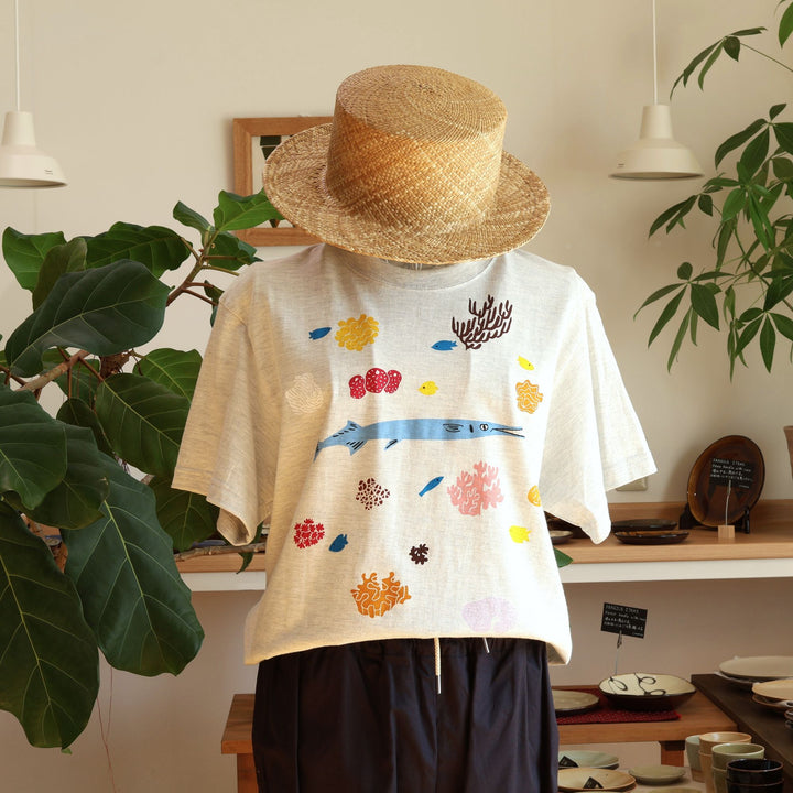 Doucatty（ドゥカティ）の手捺染Tシャツ「ダツくん」。ナチュラルなオートミール（またはライトグレー）地に、鮮やかな水色で描かれたダツを中心に、黄色やピンク、ブラウンのカラフルなサンゴや海中植物が全体に散りばめられた華やかな手刷りプリント。沖縄製。