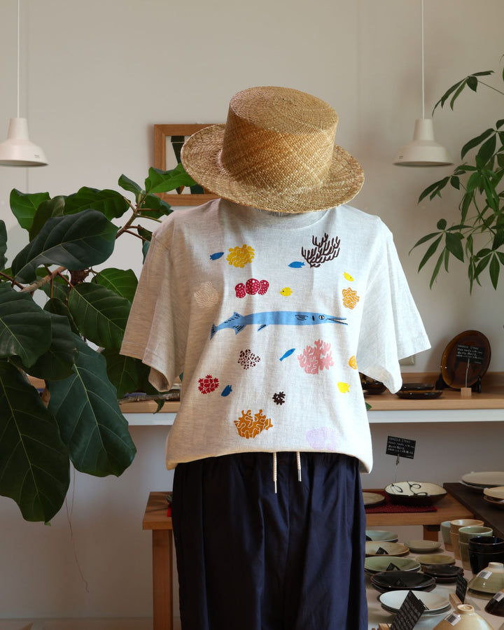 Doucatty（ドゥカティ）の手捺染Tシャツ「ダツくん」。ナチュラルなオートミール（またはライトグレー）地に、鮮やかな水色で描かれたダツを中心に、黄色やピンク、ブラウンのカラフルなサンゴや海中植物が全体に散りばめられた華やかな手刷りプリント。沖縄製。