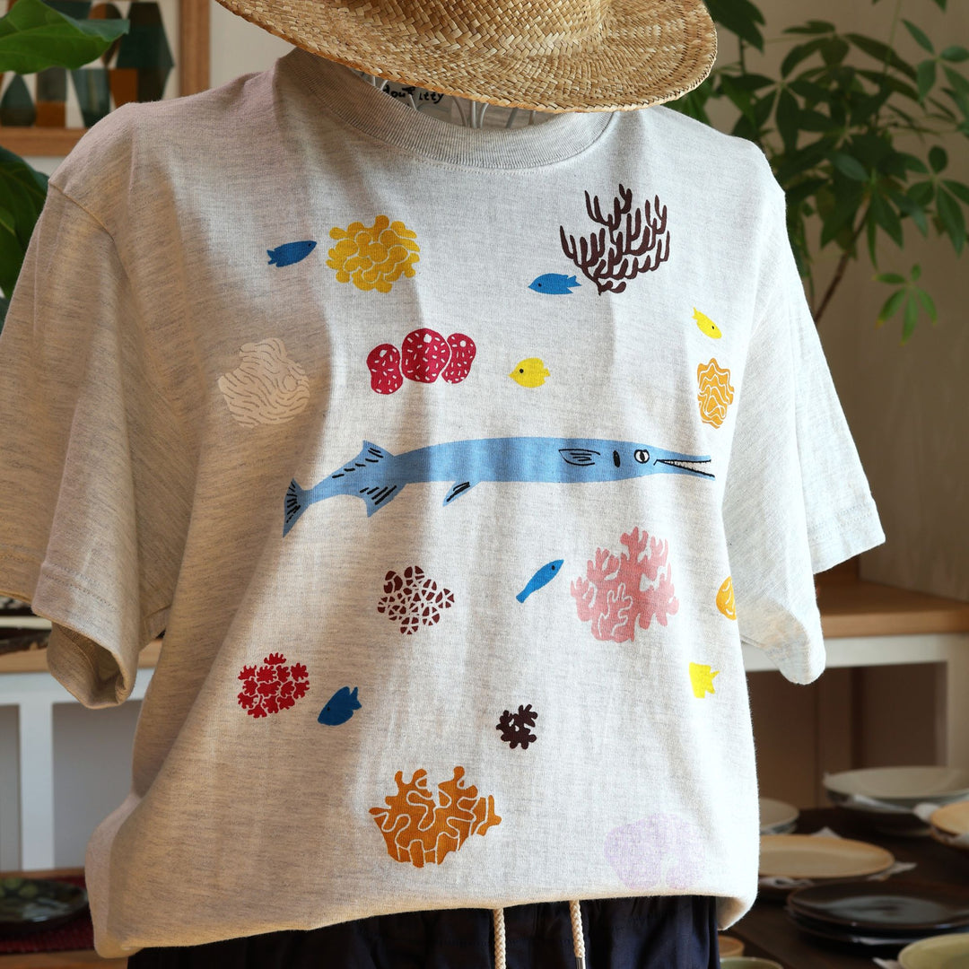 Doucatty（ドゥカティ）の手捺染Tシャツ「ダツくん」。ナチュラルなオートミール（またはライトグレー）地に、鮮やかな水色で描かれたダツを中心に、黄色やピンク、ブラウンのカラフルなサンゴや海中植物が全体に散りばめられた華やかな手刷りプリント。沖縄製。