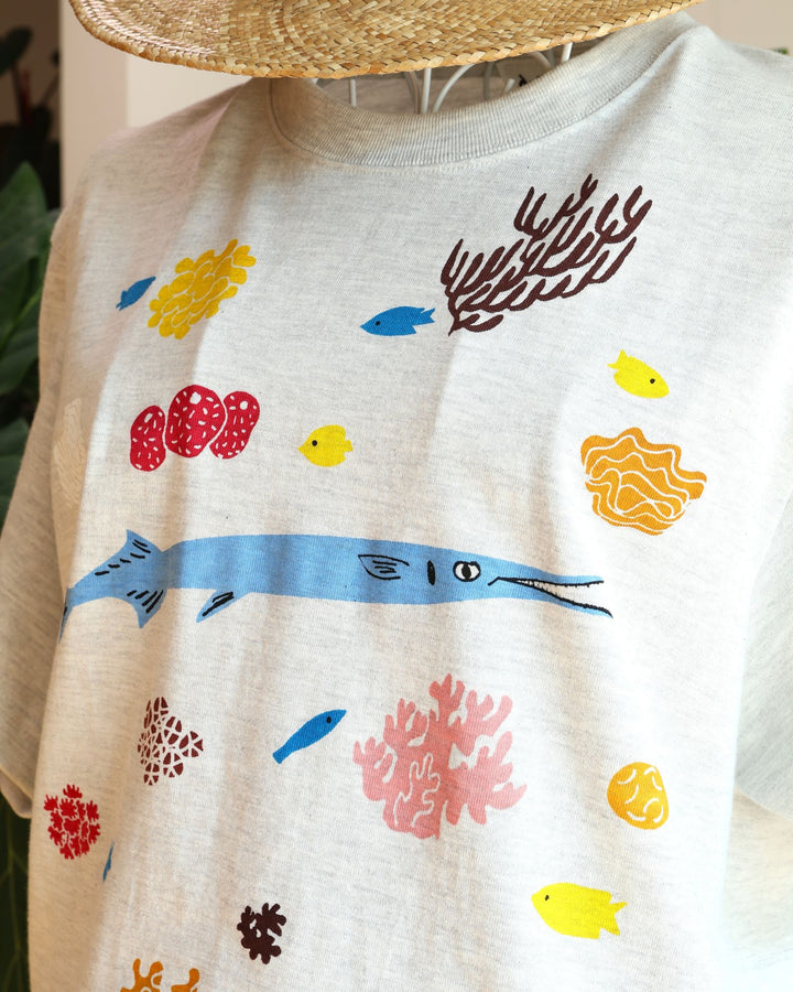 Doucatty（ドゥカティ）の手捺染Tシャツ「ダツくん」。ナチュラルなオートミール（またはライトグレー）地に、鮮やかな水色で描かれたダツを中心に、黄色やピンク、ブラウンのカラフルなサンゴや海中植物が全体に散りばめられた華やかな手刷りプリント。沖縄製。