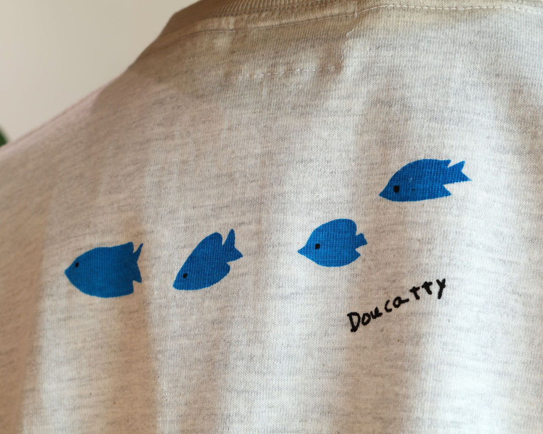 Doucatty（ドゥカティ）の手捺染Tシャツ「ダツくん」の背面。肩のあたりに、鮮やかな青色で描かれた小さな魚3匹が右に向かって並んで泳ぐ姿と「Doucatty」の手書きロゴがプリントされている。沖縄製。