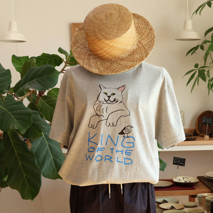 沖縄の工房、Doucatty（ドゥカティ）の手捺染Tシャツ「King of The World」。杢グレー地に、前脚を組んで不敵な笑みを浮かべる堂々とした猫と、手描き青文字の「KING OF THE WORLD」がフロント中央に描かれている。