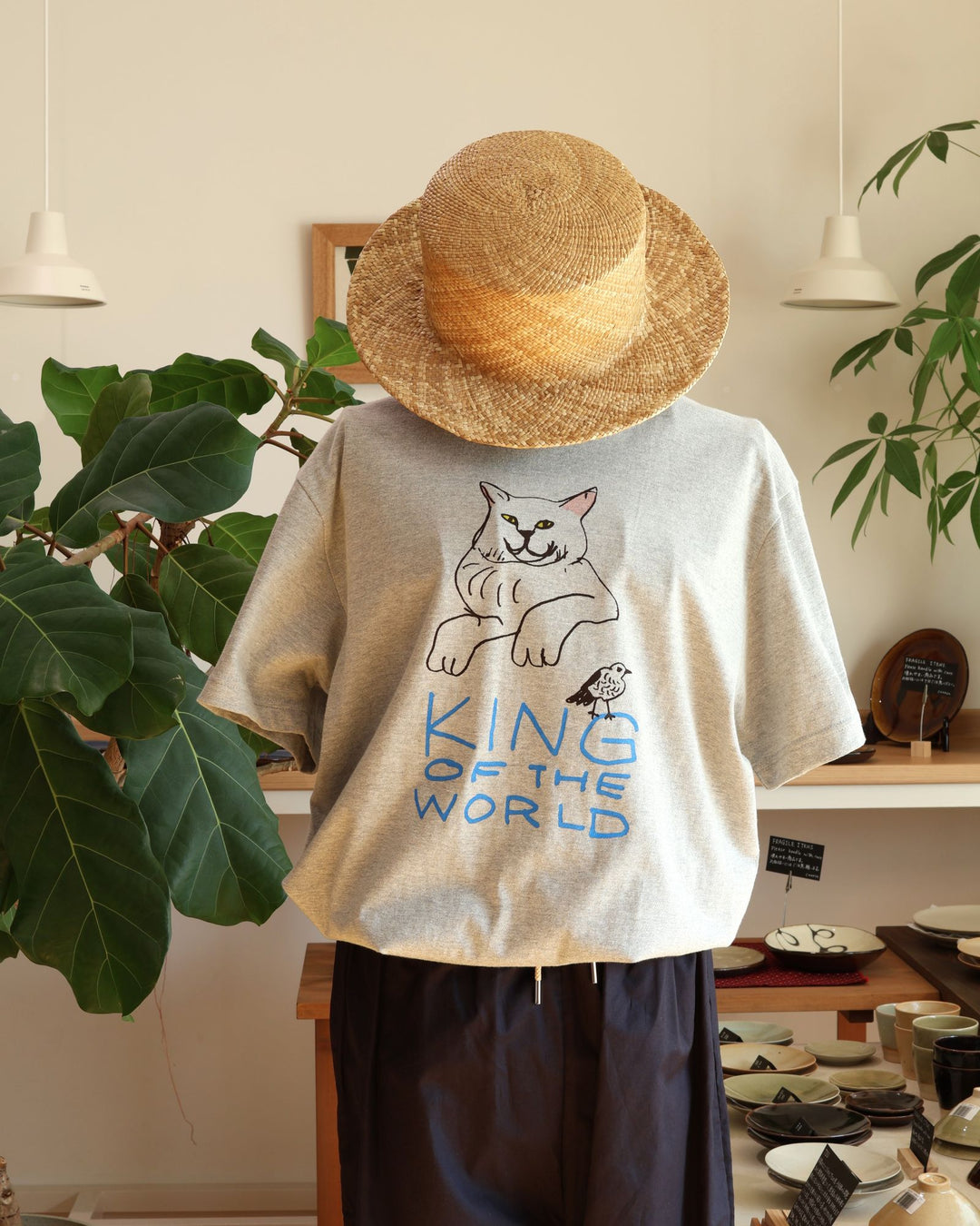 沖縄の工房、Doucatty（ドゥカティ）の手捺染Tシャツ「King of The World」。杢グレー地に、前脚を組んで不敵な笑みを浮かべる堂々とした猫と、手描き青文字の「KING OF THE WORLD」がフロント中央に描かれている。