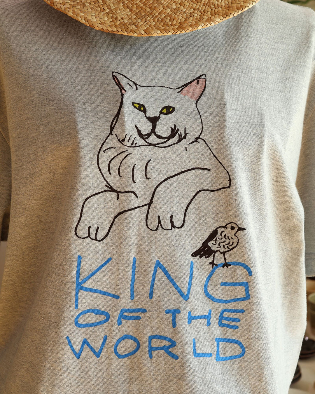 沖縄の工房、Doucatty（ドゥカティ）の手捺染Tシャツ「King of The World」。杢グレー地に、前脚を組んで不敵な笑みを浮かべる堂々とした猫と、手描き青文字の「KING OF THE WORLD」がフロント中央に描かれている。