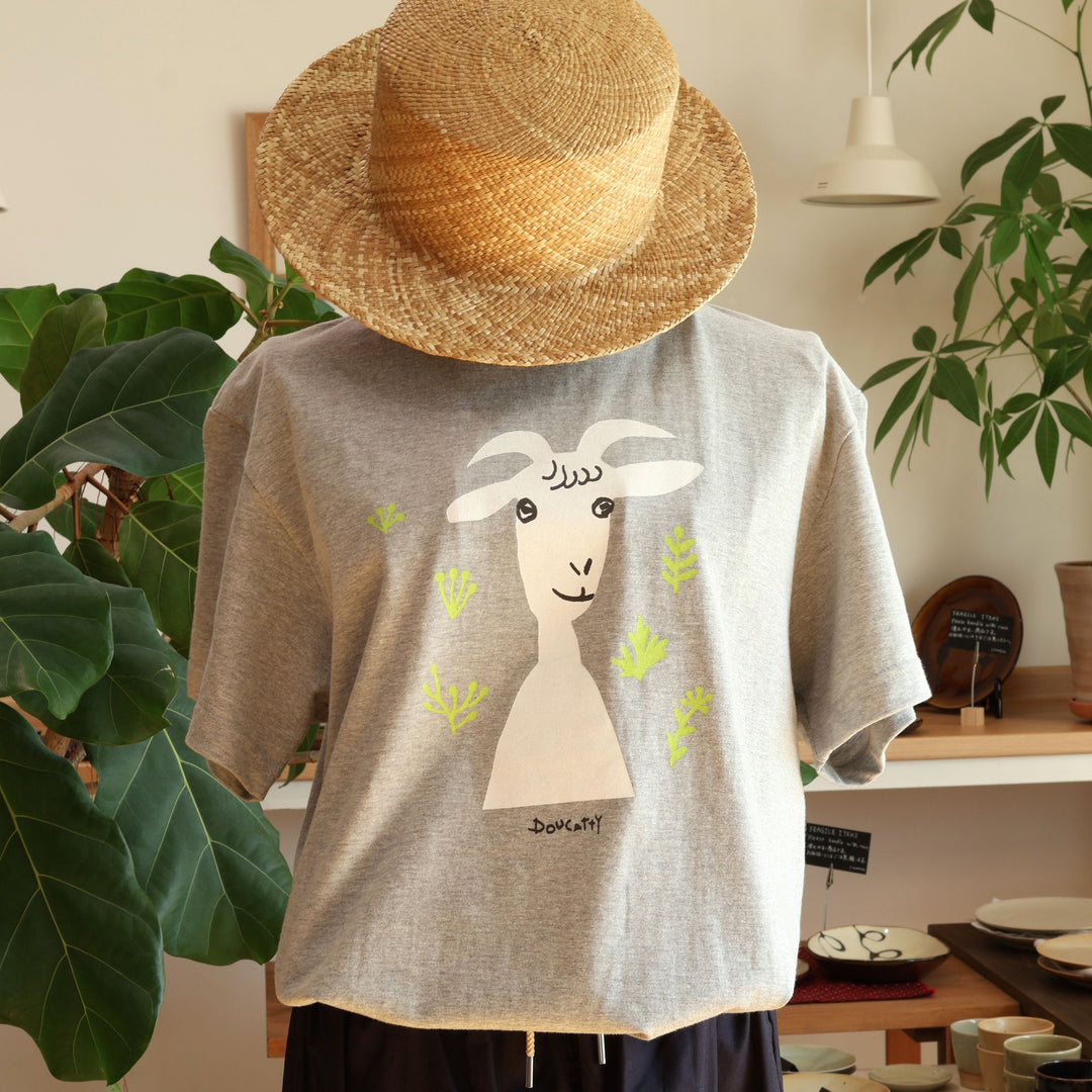 Doucatty（ドゥカティ）の手捺染Tシャツ「ヤギのメリー」。落ち着いた杢グレー地に、白い顔とひょうきんな目元が愛らしいヤギの「メリー」がフロント中央に描かれ、首元には鮮やかな黄緑色の草花があしらわれている。