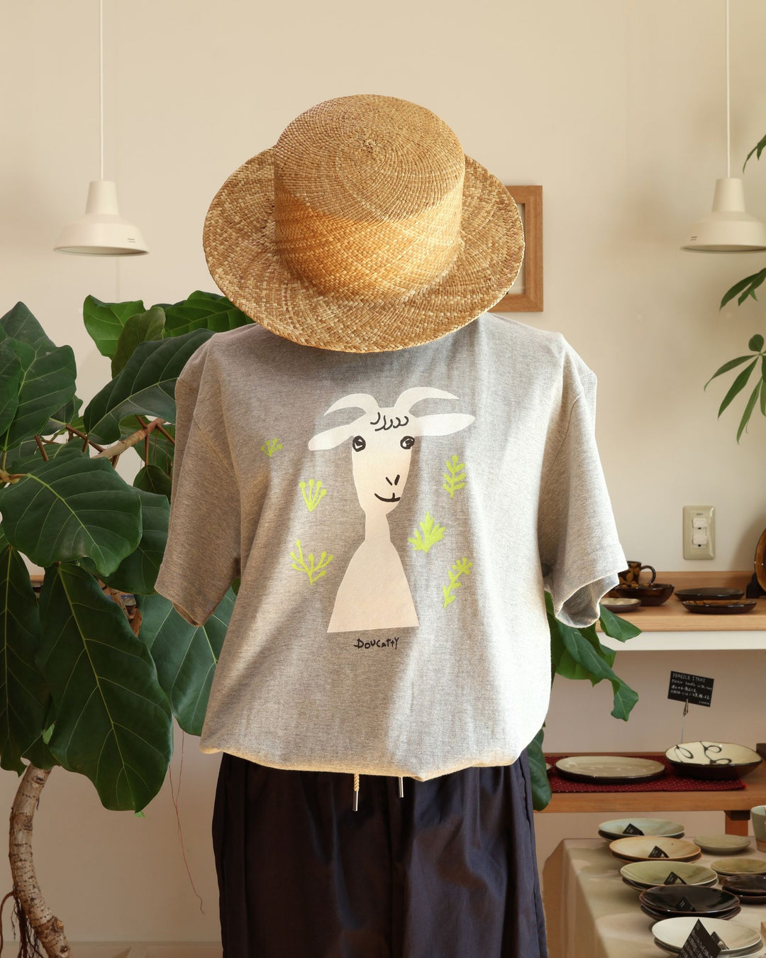 Doucatty（ドゥカティ）の手捺染Tシャツ「ヤギのメリー」。落ち着いた杢グレー地に、白い顔とひょうきんな目元が愛らしいヤギの「メリー」がフロント中央に描かれ、首元には鮮やかな黄緑色の草花があしらわれている。