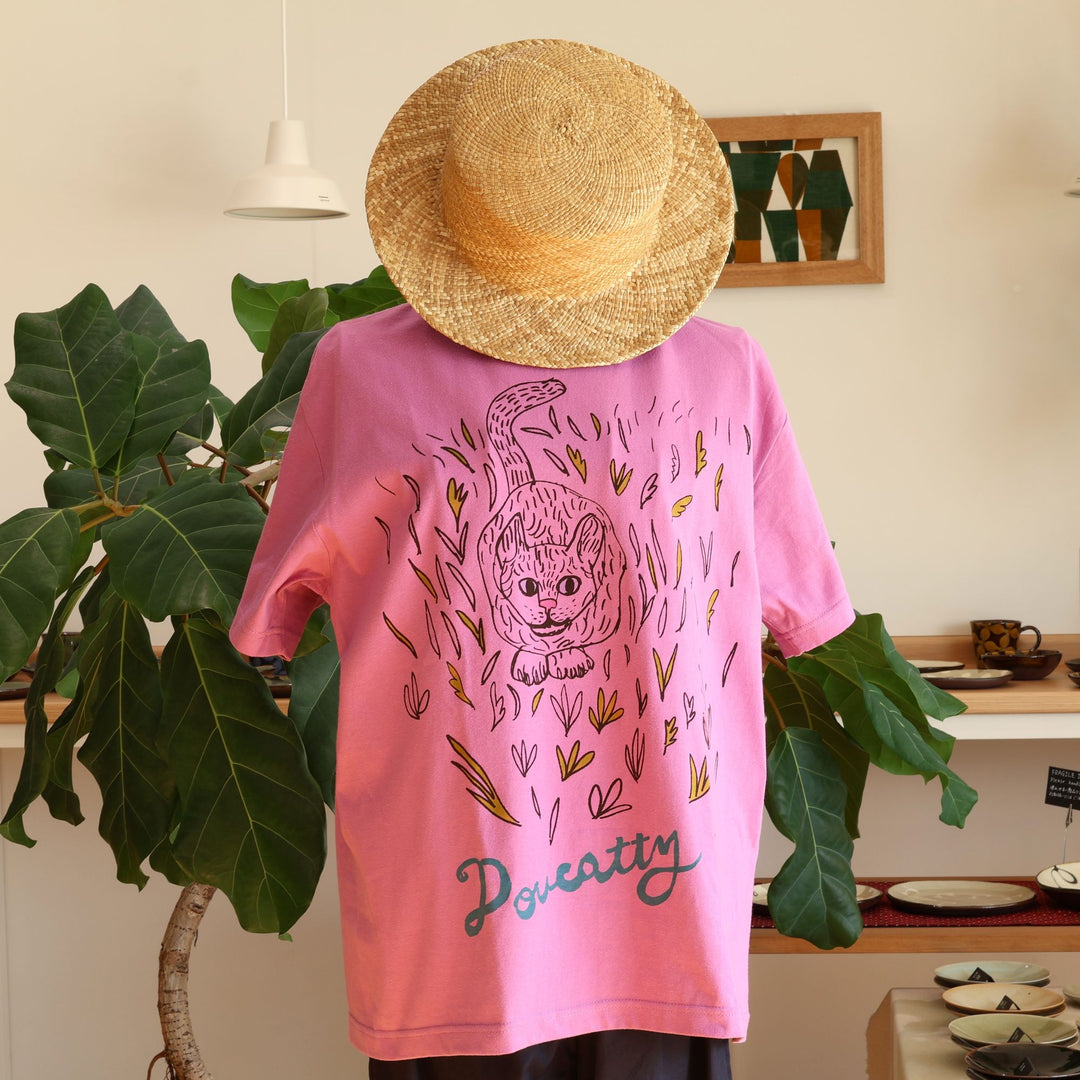 沖縄 Doucatty（ドゥカティ）の手捺染Tシャツ「LAVY」。鮮やかなあざみピンク地のバックプリントに、草むらから愛らしく顔をのぞかせる猫「LAVY」の繊細な黒線画イラストと「Doucatty」のロゴがデザインされている。