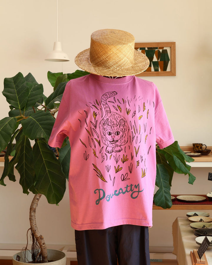 沖縄 Doucatty（ドゥカティ）の手捺染Tシャツ「LAVY」。鮮やかなあざみピンク地のバックプリントに、草むらから愛らしく顔をのぞかせる猫「LAVY」の繊細な黒線画イラストと「Doucatty」のロゴがデザインされている。