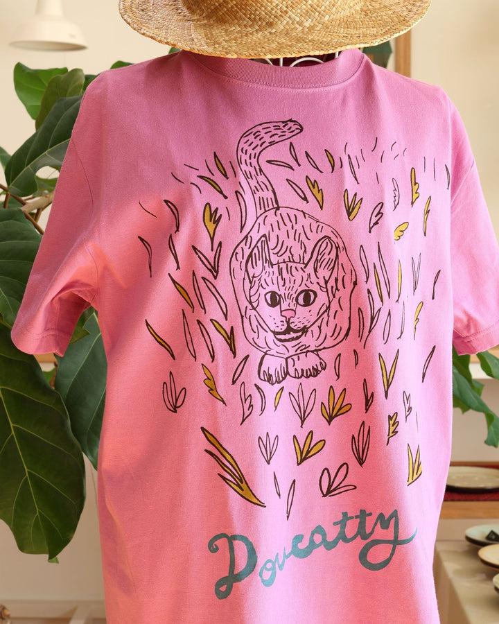 沖縄 Doucatty（ドゥカティ）の手捺染Tシャツ「LAVY」。鮮やかなあざみピンク地のバックプリントに、草むらから愛らしく顔をのぞかせる猫「LAVY」の繊細な黒線画イラストと「Doucatty」のロゴがデザインされている。