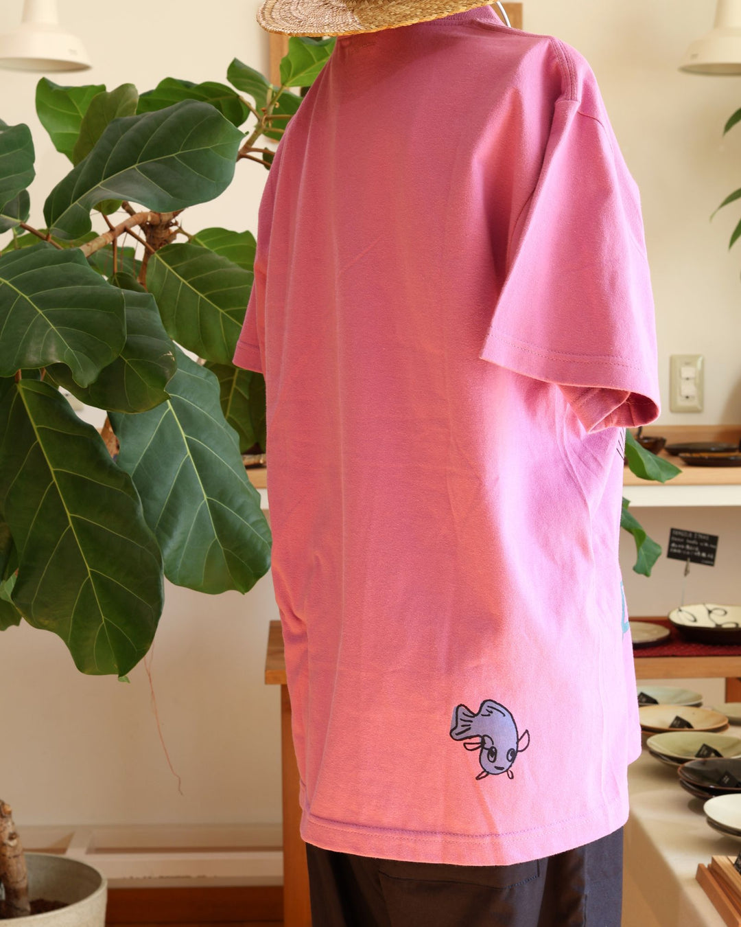 沖縄 Doucatty（ドゥカティ）の手捺染Tシャツ「LAVY」。鮮やかなあざみピンク地のバックプリントに、草むらから愛らしく顔をのぞかせる猫「LAVY」の繊細な黒線画イラストと「Doucatty」のロゴがデザインされている。