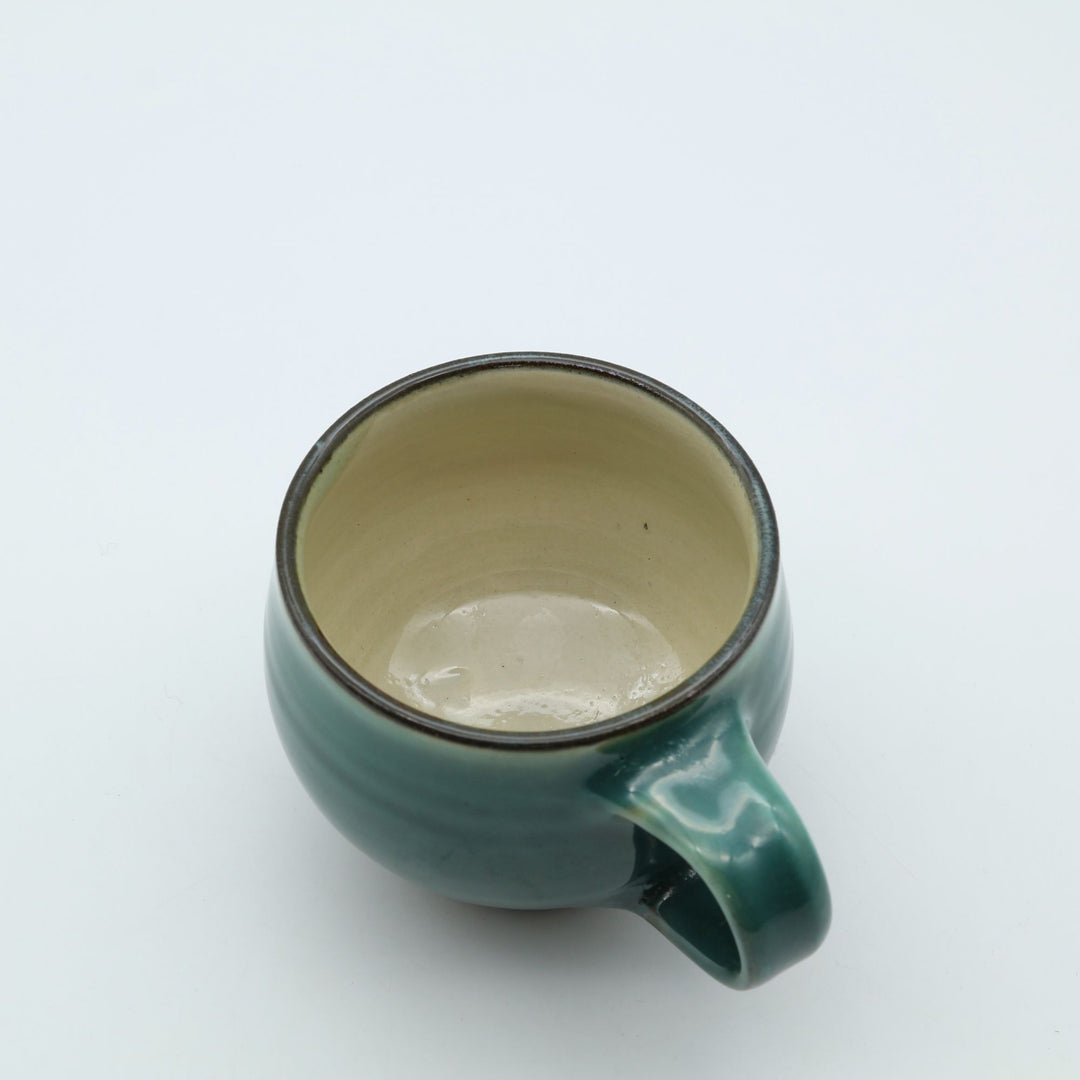大分県の伝統工芸・小鹿田焼の緑釉（青流し）が美しい陶器のマグカップ。手でしっかりと幅広の取っ手を持ち、サイズ感やコロンとした丸みのあるフォルム。内側はクリーミーな白釉の暮らしの器。canosa取扱商品。