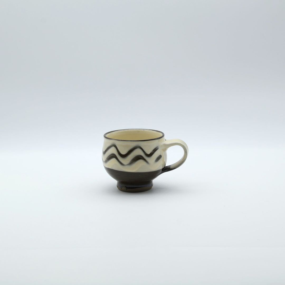 大分県の伝統工芸・小鹿田焼の指描き模様が施された陶器のマグカップ。白釉をベースに黒い波状の指描きがリズミカルに描かれ、底面がブラウンのコーヒーカラーに切り替わった、幅広の取っ手が特徴的な暮らしの器。canosa取扱商品。