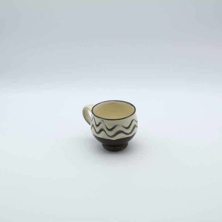 大分県の伝統工芸・小鹿田焼の指描き模様が施された陶器のマグカップ。白釉をベースに黒い波状の指描きがリズミカルに描かれ、底面がブラウンのコーヒーカラーに切り替わった、幅広の取っ手が特徴的な暮らしの器。canosa取扱商品。