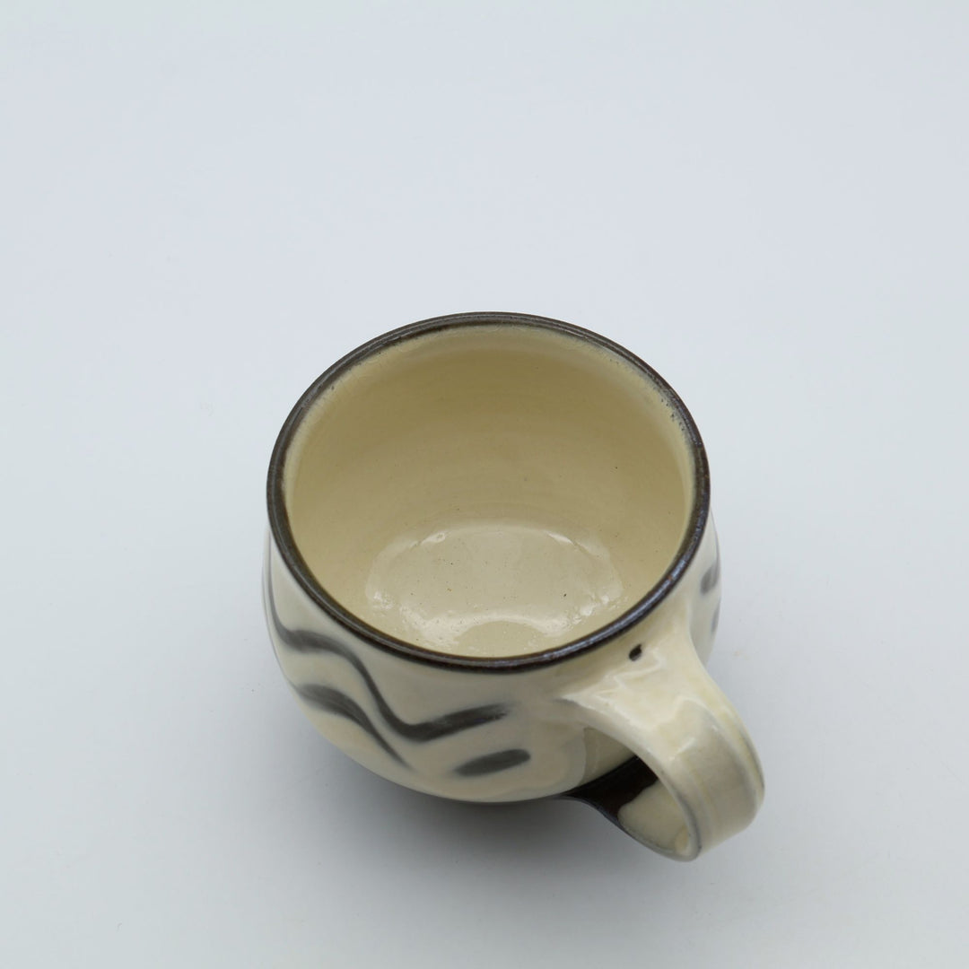 大分県の伝統工芸・小鹿田焼の指描き模様が施された陶器のマグカップ。白釉をベースに黒い波状の指描きがリズミカルに描かれ、底面がブラウンのコーヒーカラーに切り替わった、幅広の取っ手が特徴的な暮らしの器。canosa取扱商品。