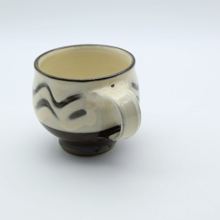大分県の伝統工芸・小鹿田焼の指描き模様が施された陶器のマグカップ。白釉をベースに黒い波状の指描きがリズミカルに描かれ、底面がブラウンのコーヒーカラーに切り替わった、幅広の取っ手が特徴的な暮らしの器。canosa取扱商品。