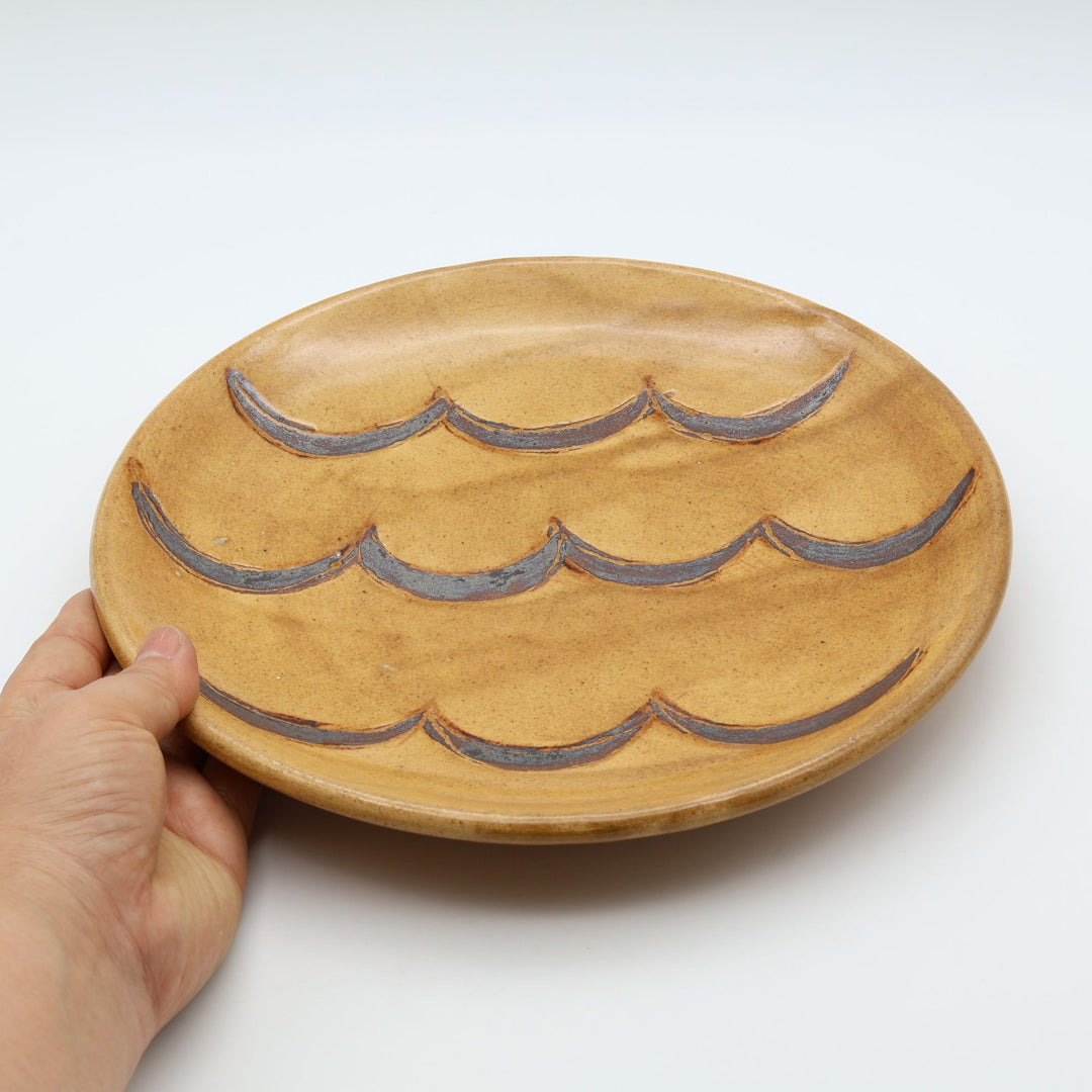 沖縄のやちむん（暮らしの器）。ベージュ・はちみつ色の優しい色合いに、波る模様が施されたシンプルな7寸皿。主菜やワンプレートに活躍する厚みのある丸皿。